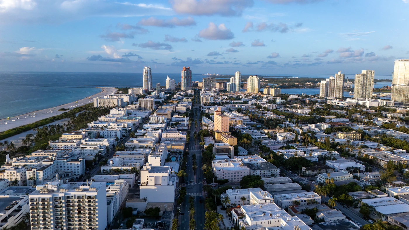 Aerial view of Miami Beach: flat white rooftops with air-conditioning units, condo towers, and the ocean under a sunny sky